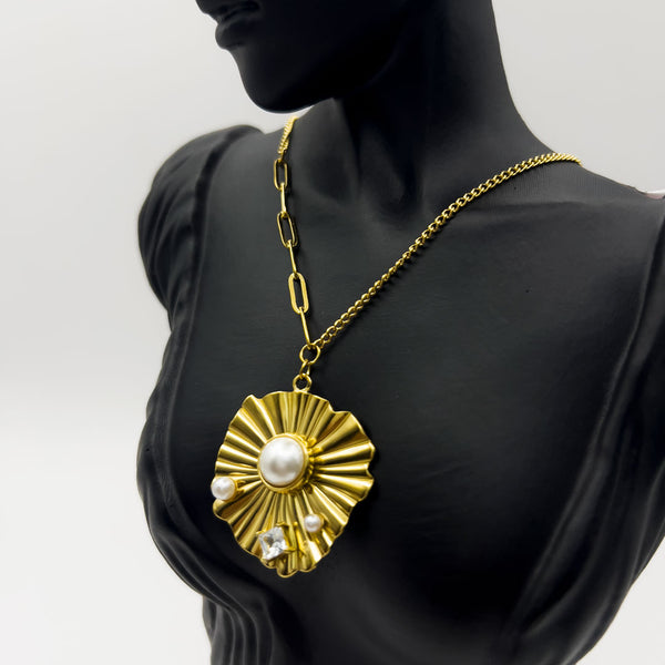 Gold necklace with a large sunburst pendant featuring pearls and crystals on a black mannequin against a white background.